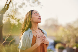 Woman standing outdoors with eyes closed and hands placed over her chest, surrounded by natural light and greenery