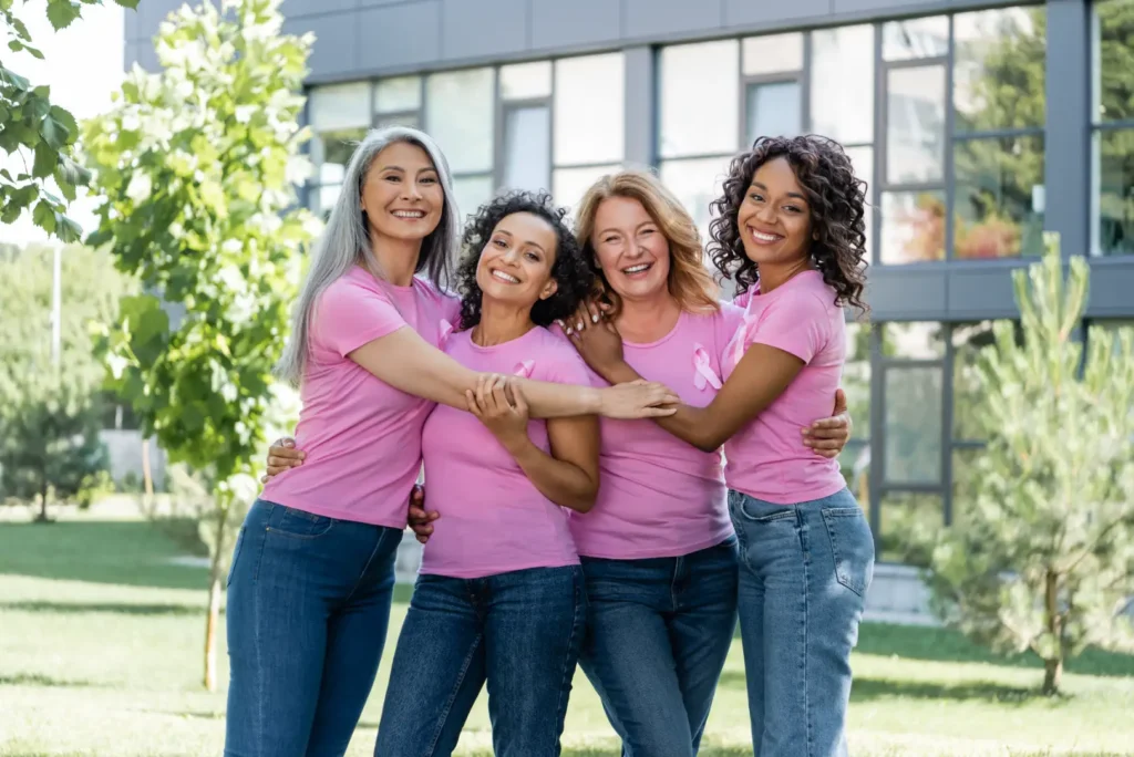 Four women in matching pink shirts with awareness ribbons stand together outdoors with their arms around one another.