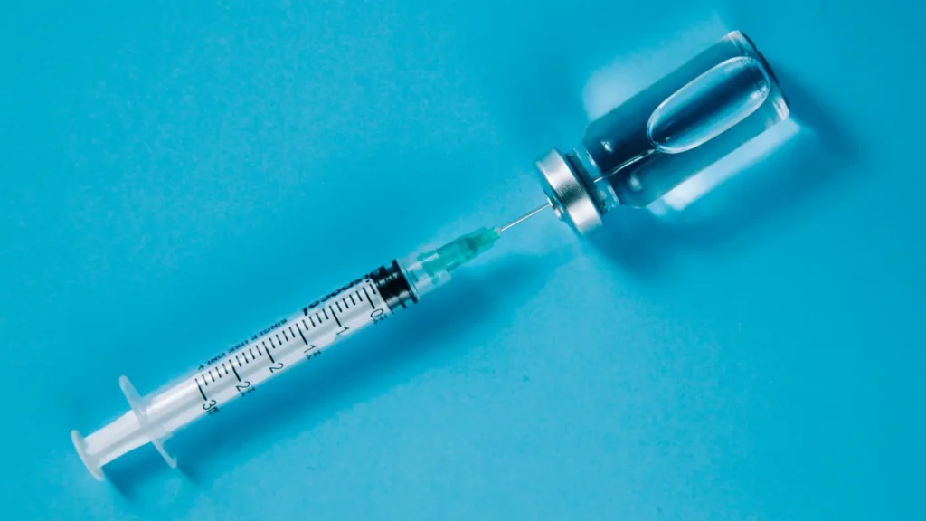 Syringe with its needle inserted into a clear glass vial on a turquoise surface.